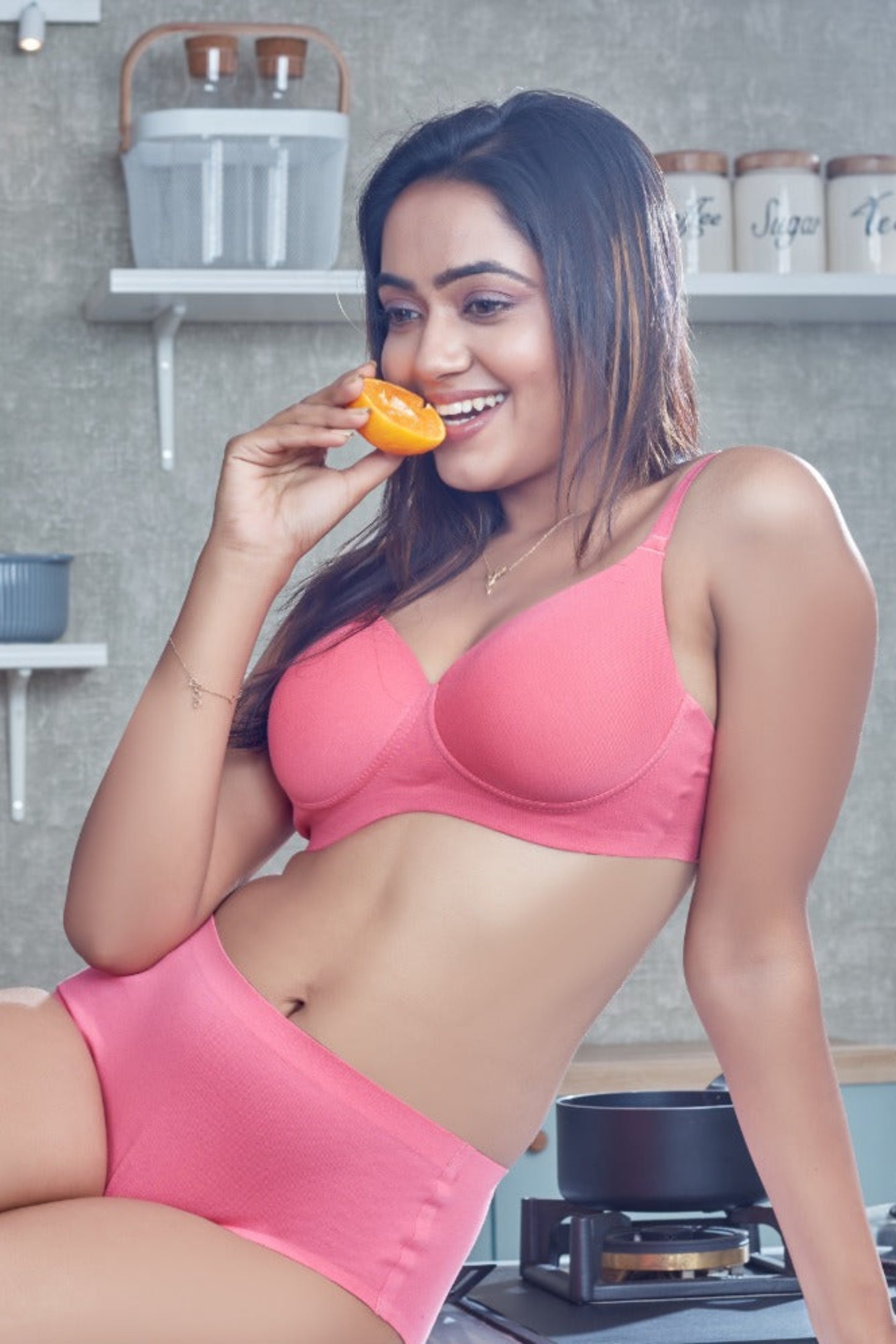 Woman in pink lingerie sitting in a kitchen holding an orange.